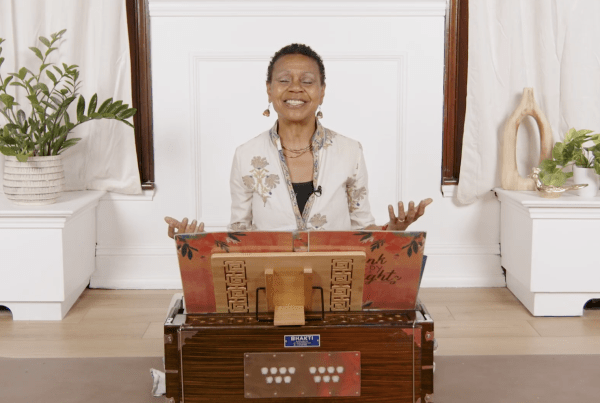 Julie Pasqual in front of a harmonium with her hands in front of her through devotion for the goddess, Radha, she is mentioning in her workshop, Three Goddesses: One Source